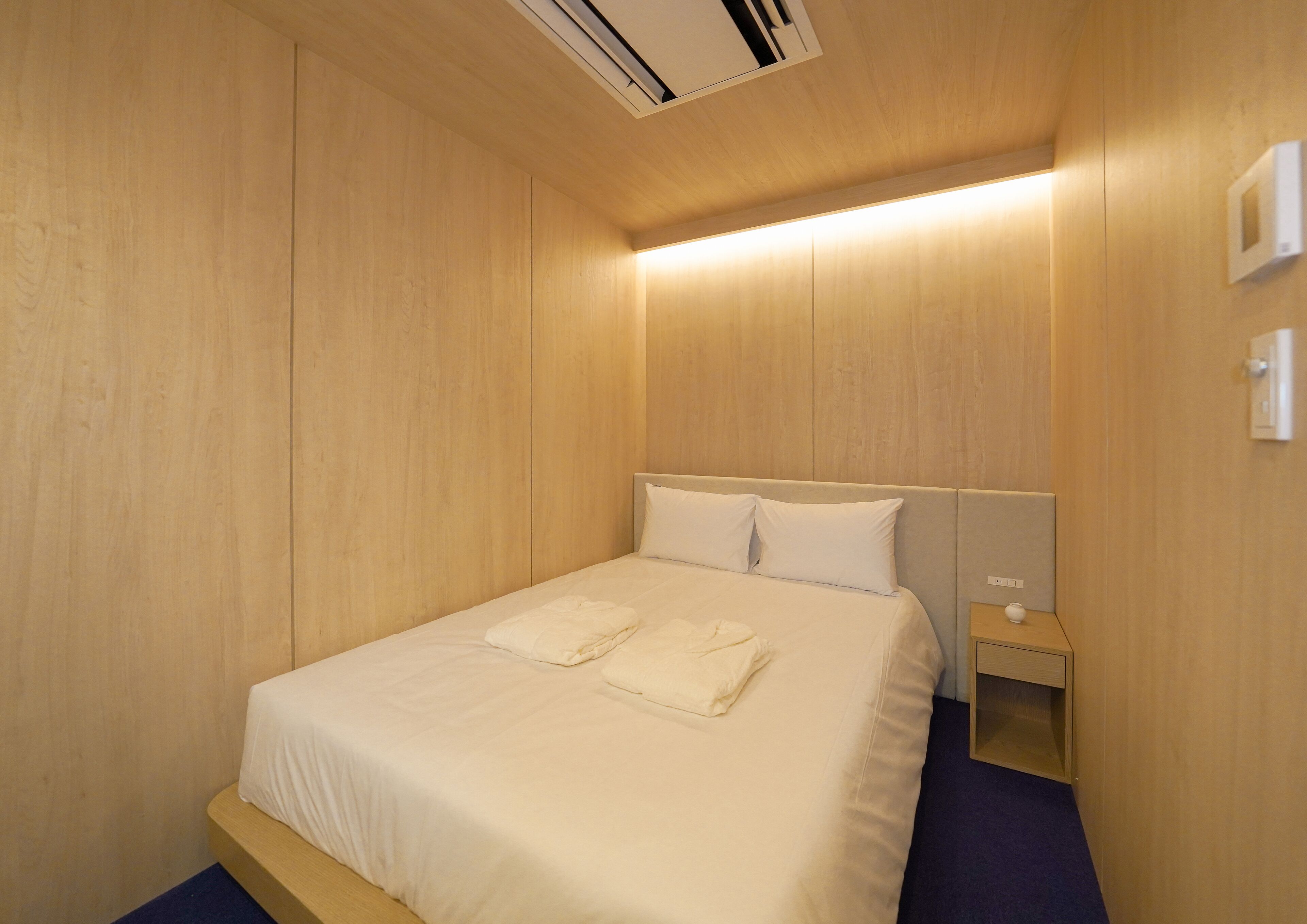 A hotel room with a wooden wall, a bed with white linens, a nightstand, and a wall-mounted air conditioner.