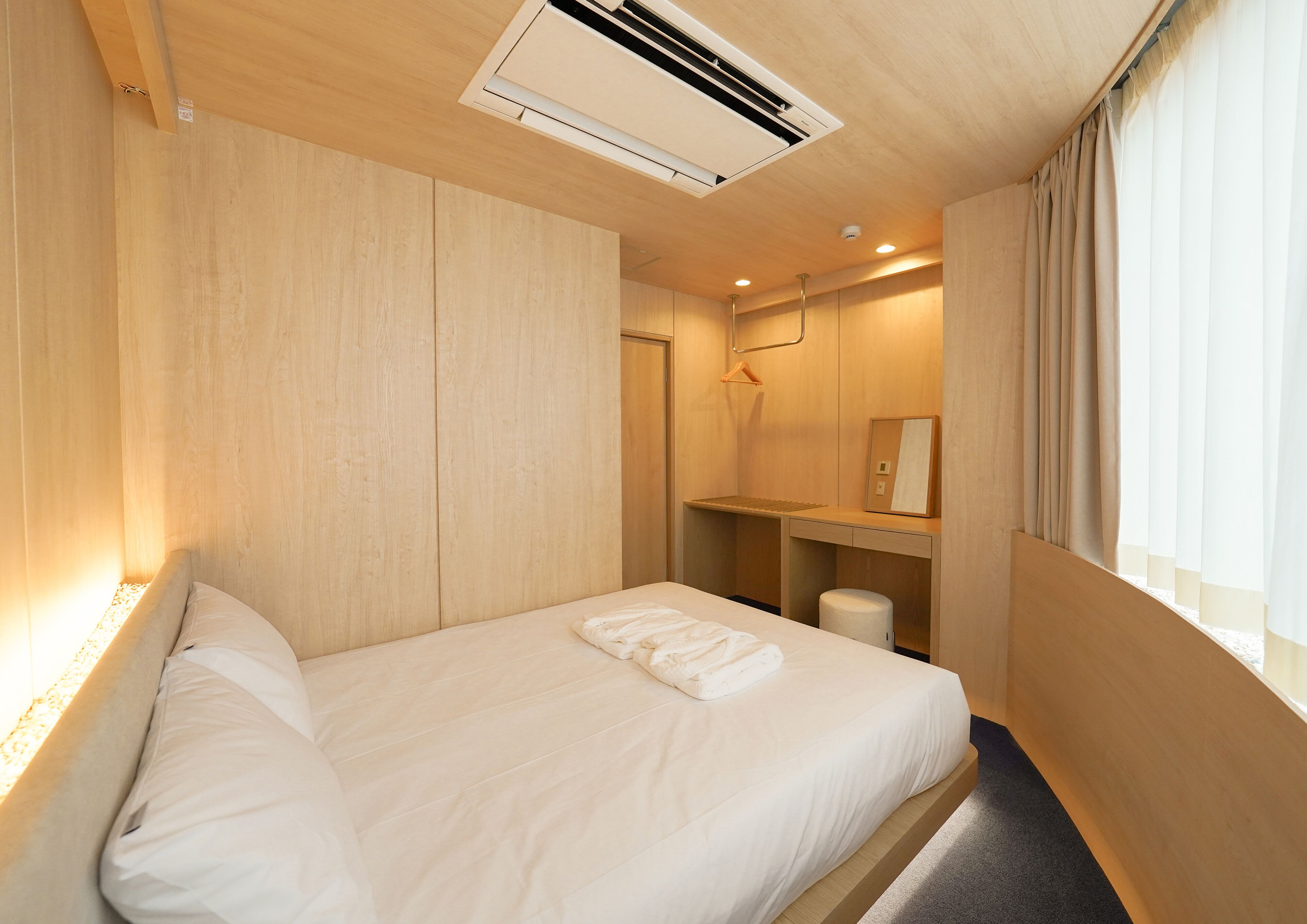Habitación de hotel con techo de madera, una cama con ropa de cama blanca, una mesita de noche pequeña, un espejo y una ventana con cortinas.