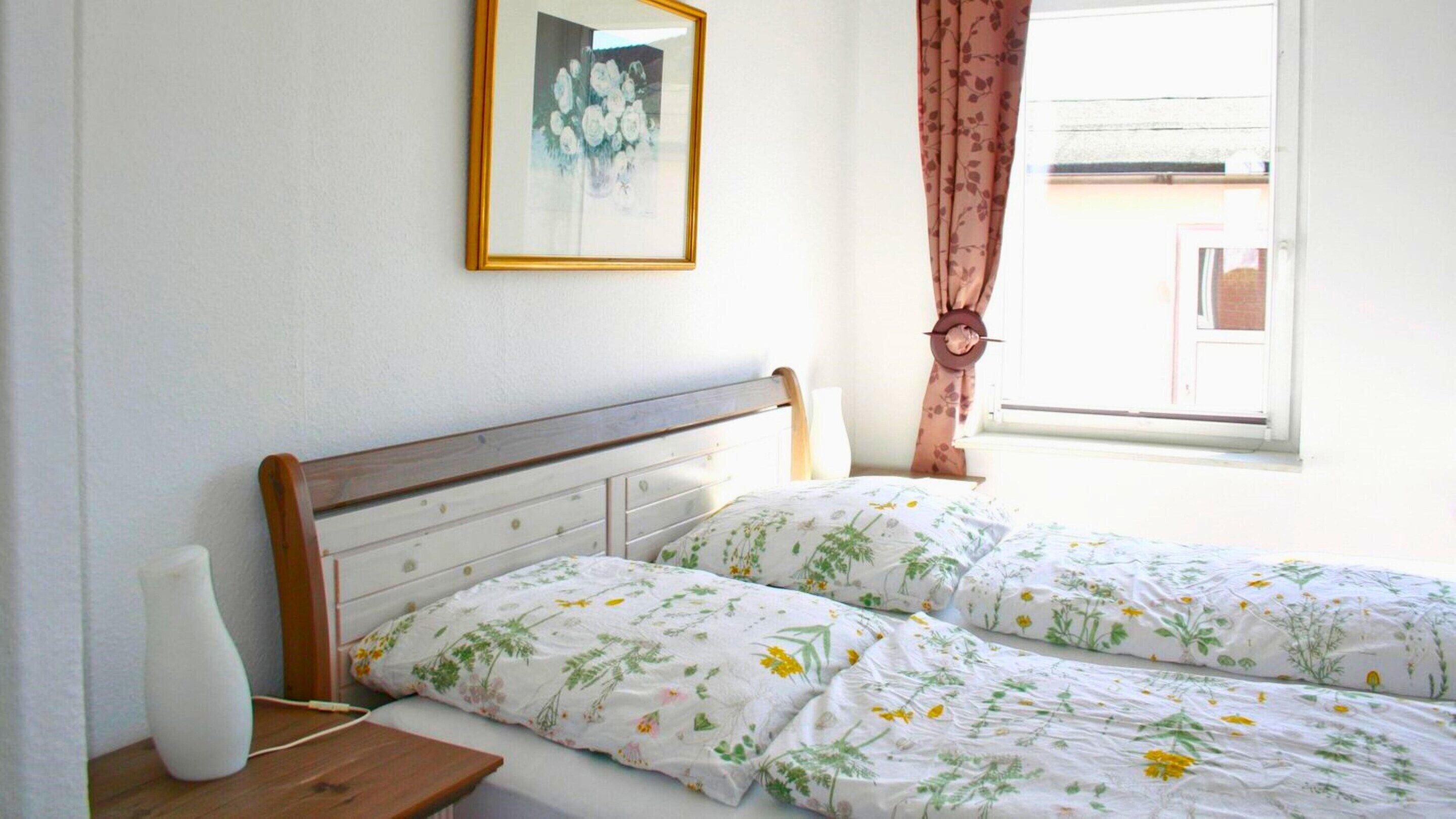 A bedroom with a wooden headboard, two beds with floral bedding, a bedside table with a lamp, and a window with curtains.