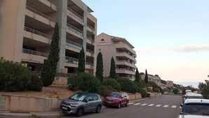 A multi-story residential building with balconies, parked cars, and a clear sky.
