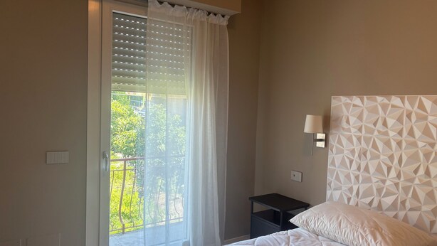 Une chambre à coucher comprenant un lit, une table de chevet, une lampe fixée au mur et une fenêtre avec des stores.