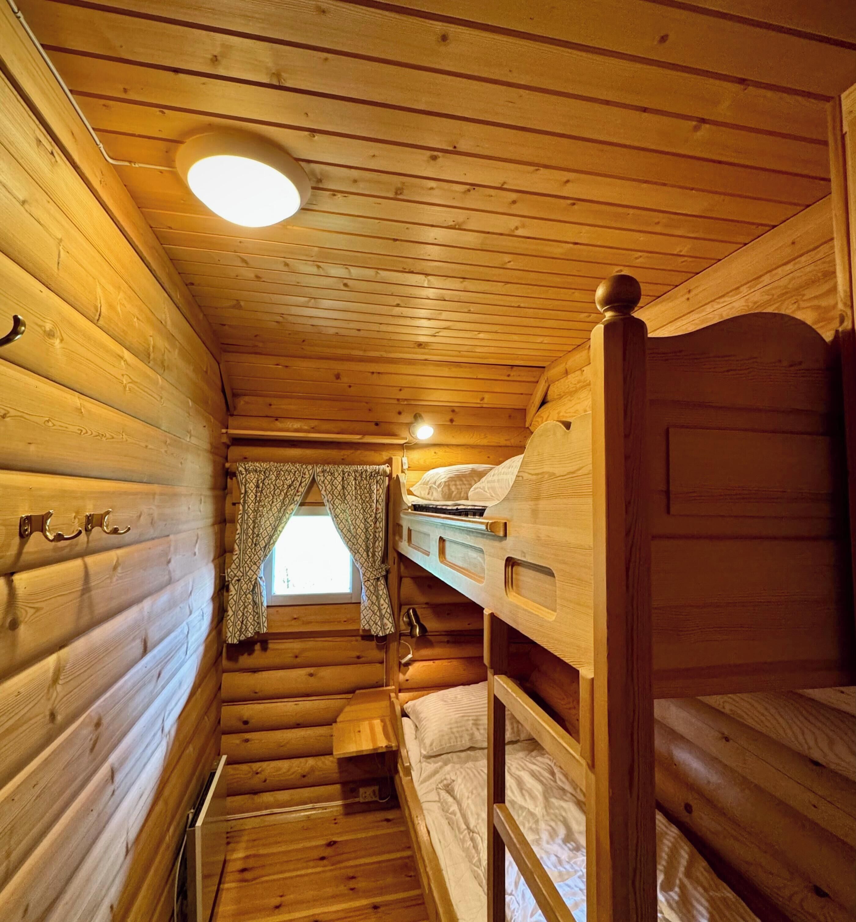 A cabin interior with wooden walls, a bunk bed, a small window with curtains, and a ceiling light.