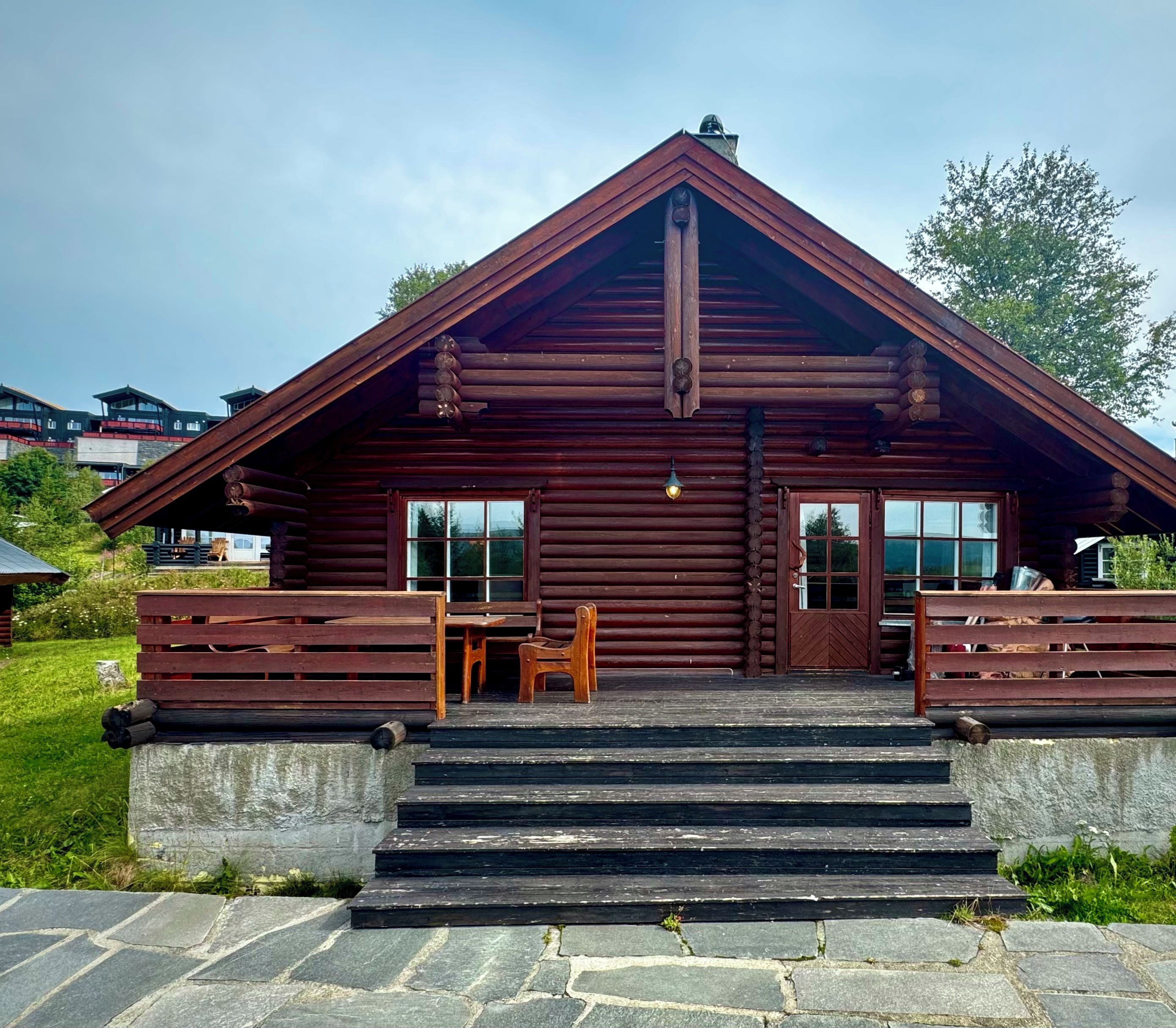 A log cabin with a steeply pitched roof, a front porch with a bench, and a stone staircase leading to the entrance.