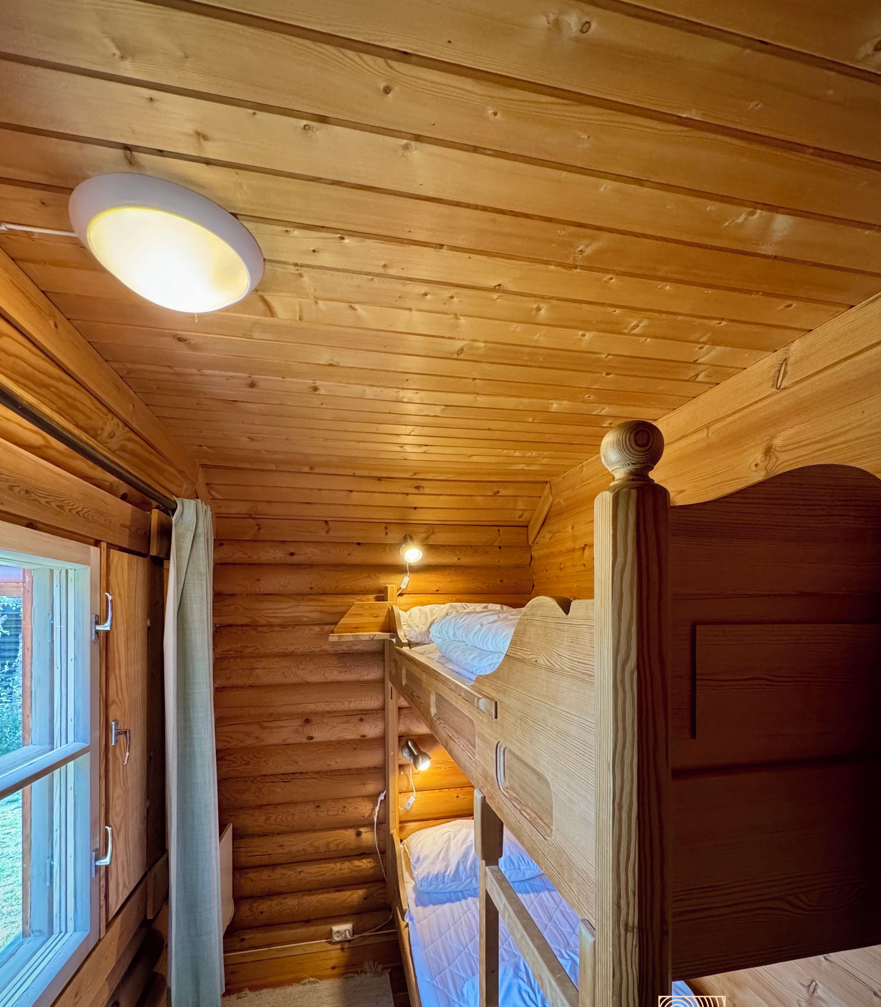 L’intérieur d’une cabine avec des murs en bois, un lit superposé, une fenêtre avec des rideaux et un luminaire au plafond.