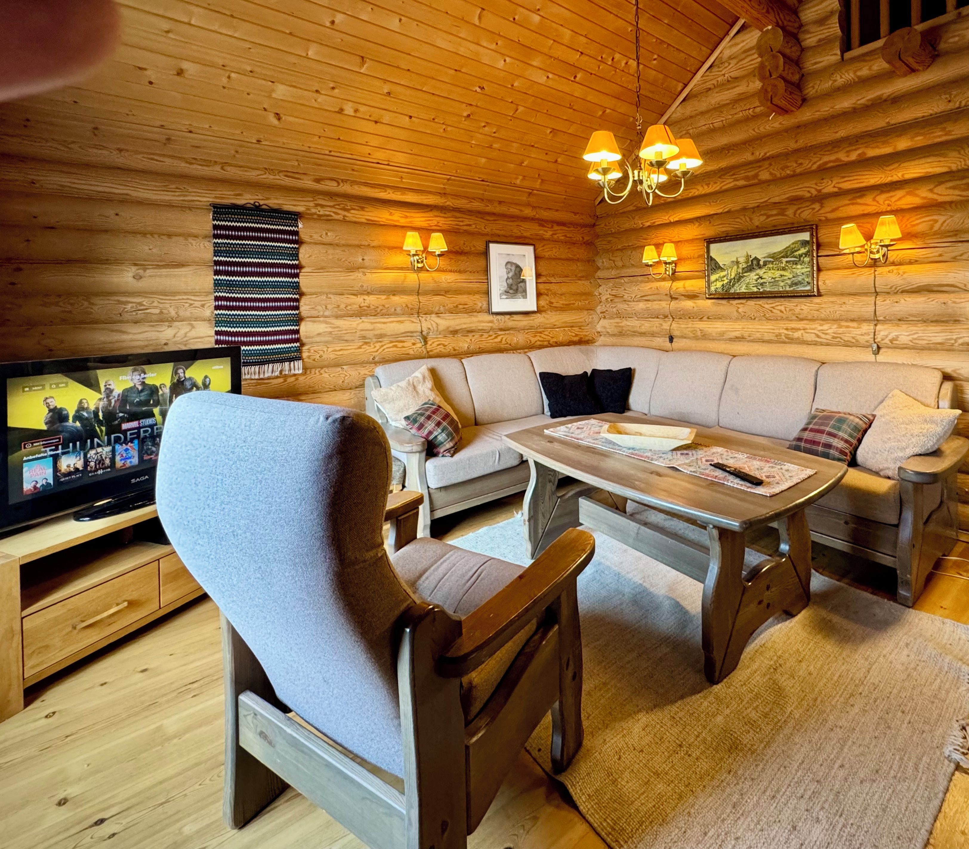 A cozy cabin interior with a wooden log wall, a sofa, a dining table, and a television displaying a movie.