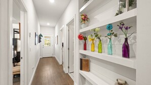 Un pasillo con paredes blancas, piso de madera y un estante blanco con jarrones de colores y una vela.