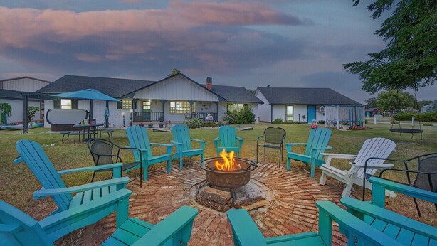 A patio area with a fire pit surrounded by Adirondack chairs, a brick fire pit, and a view of a row of buildings in the background.