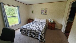 Un dormitorio con cama, dos mesitas de noche, una silla, ventana con cortinas y un cuadro en la pared.