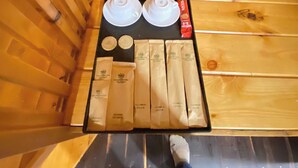 Interior de una cabaña de madera con suelo de madera, una mesa negra con dos tazas blancas, un paquete rojo y varias bolsas color beige.