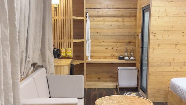 Una habitación acogedora con suelo de madera, un reposapiés redondo de mimbre, un sofá blanco, una mesita con una lámpara y una ventana con vistas al exterior.