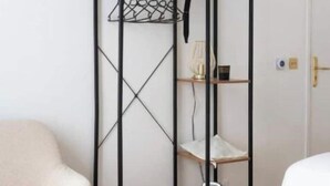 A room with a wooden floor, a white chair, a bed, and a black metal wardrobe with shelves. - Apt Tour Eiffel invalides (Paris)