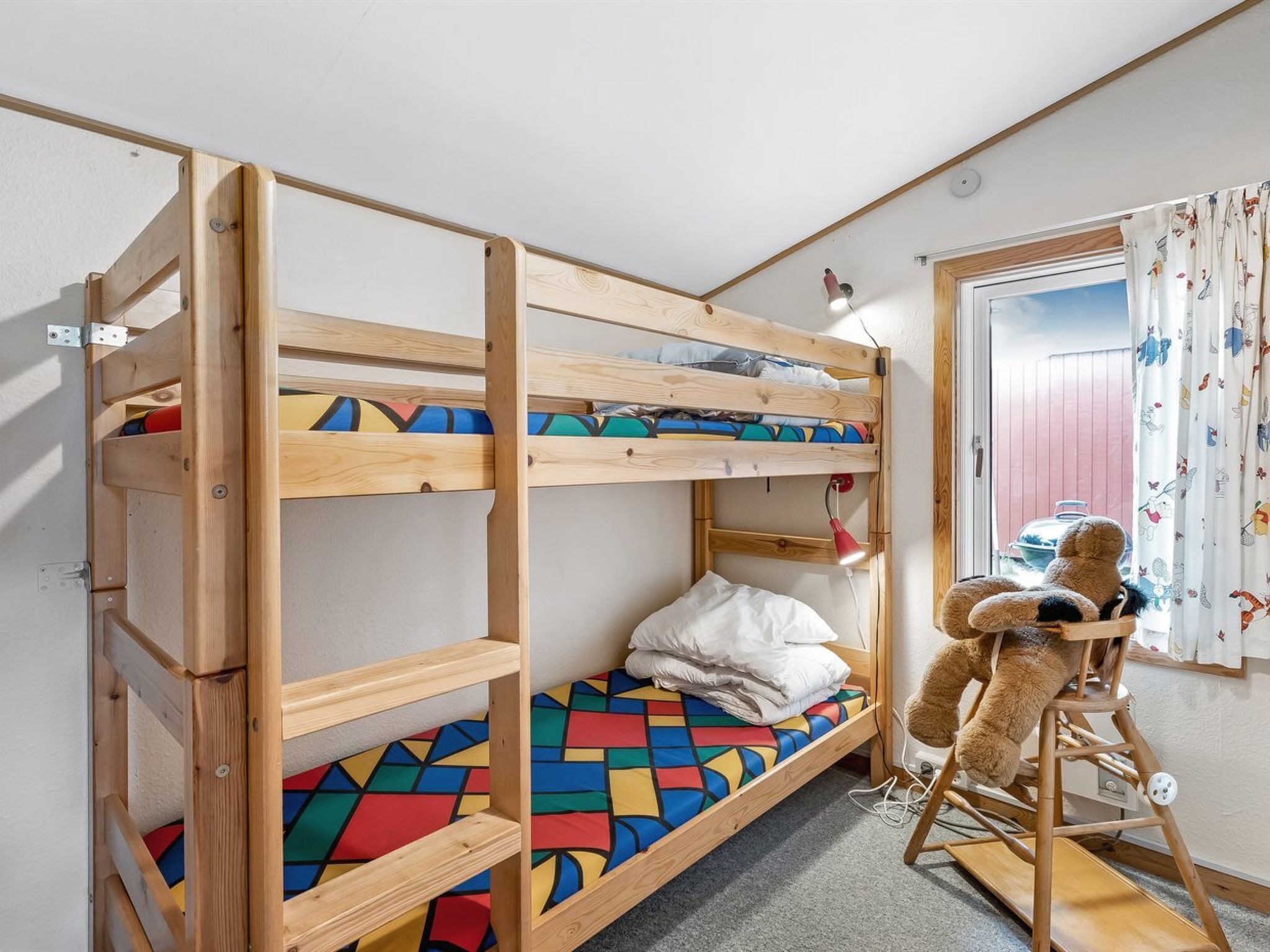 A bunk bed with a colorful geometric-patterned mattress, a small desk with a lamp, and a teddy bear on a chair.