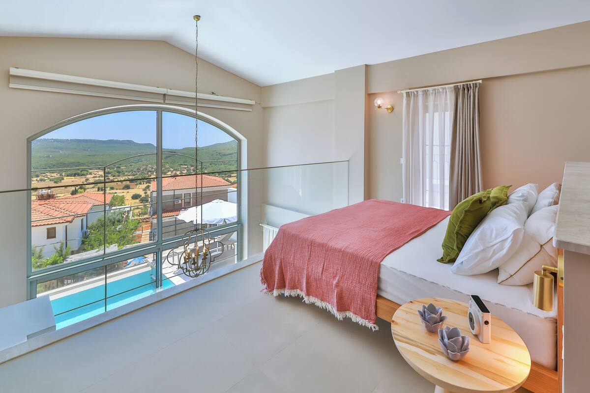 A bedroom with a large window, a bed with pillows, a small table with a speaker, and a view of buildings and hills.