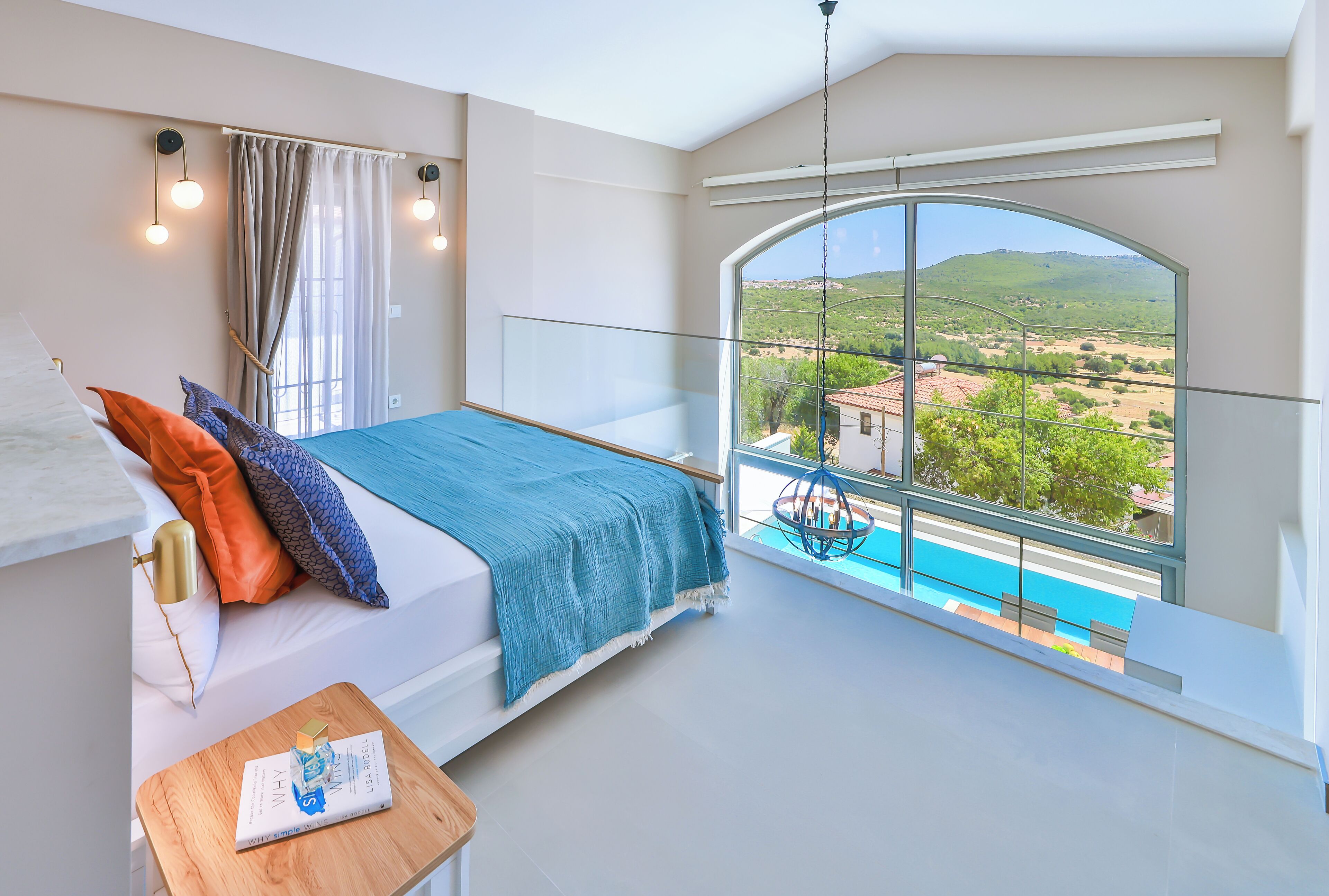 A bedroom with a large window, a bed, a bedside table, and a view of a swimming pool and landscape.