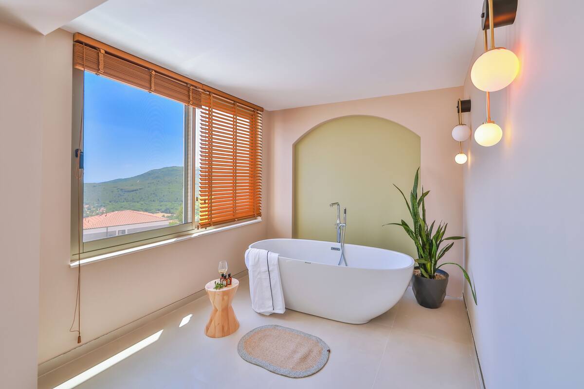 A modern bathroom with a freestanding bathtub, a large window with blinds, a potted plant, and a small wooden stool.