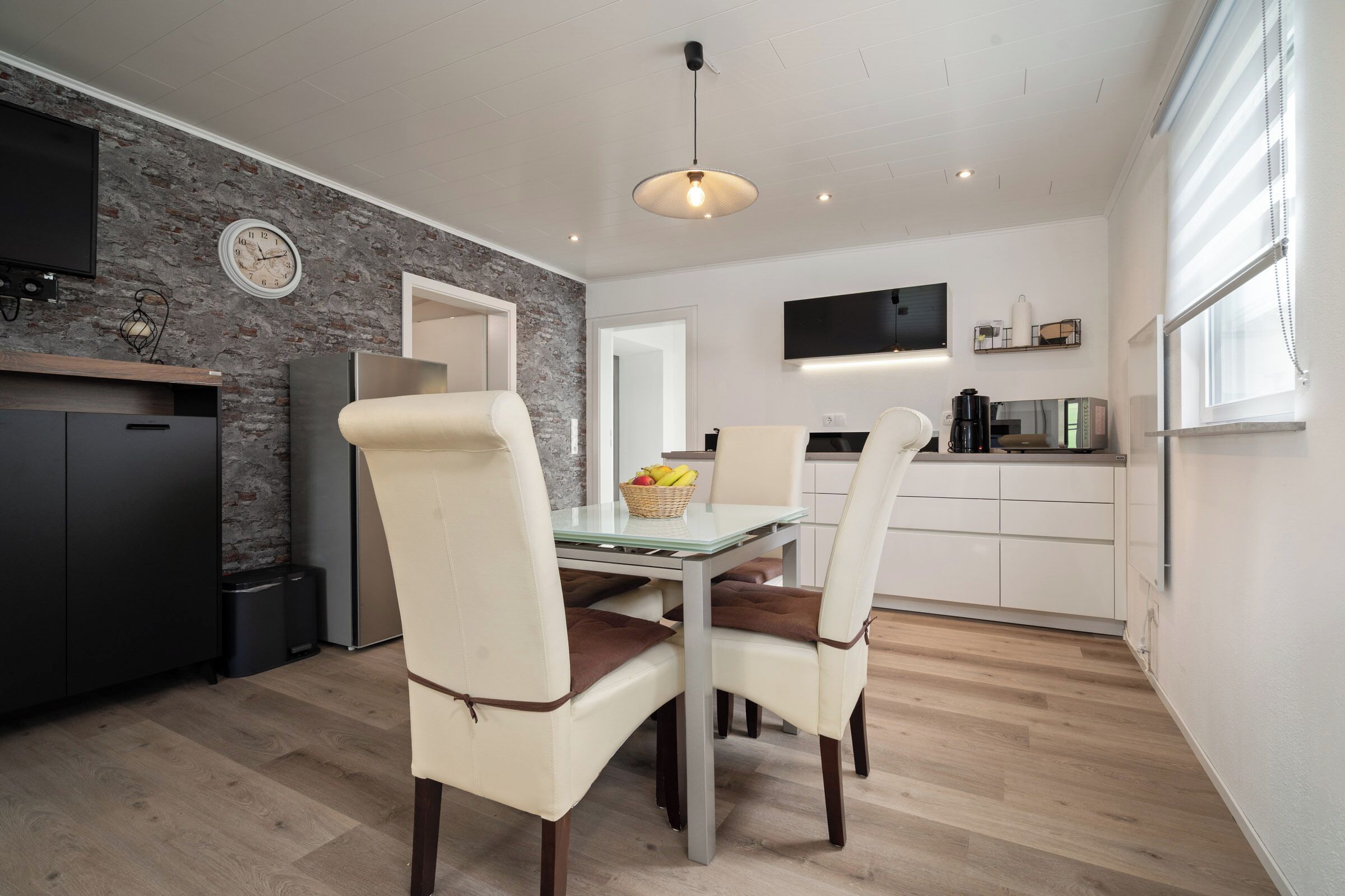 A modern kitchen with a stone wall backsplash, a dining area with two white chairs and a glass table, and a TV mounted above the counter.