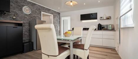 A modern kitchen with a stone wall backsplash, a dining area with two white chairs and a glass table, and a TV mounted above the counter.