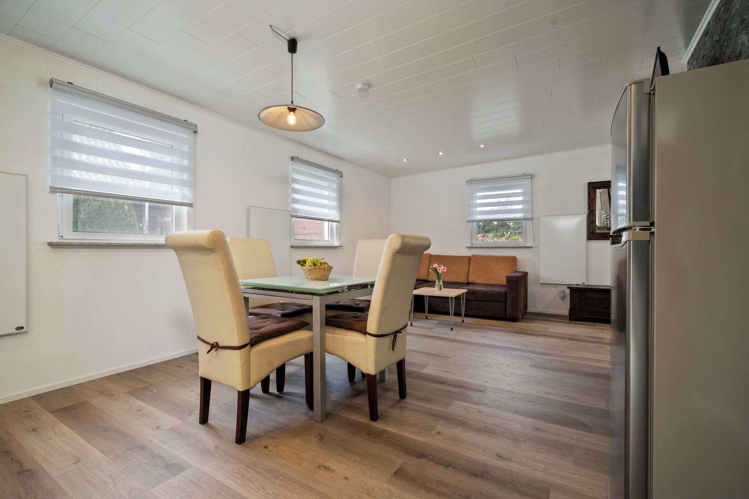 A modern kitchen with a dining area, wooden flooring, and large windows with blinds.