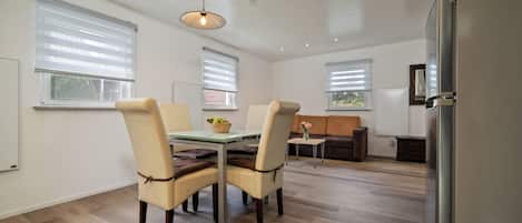 A modern kitchen with a dining area, wooden flooring, and large windows with blinds.