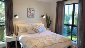 Une chambre à coucher avec un grand lit, deux lampes de chevet, une fenêtre avec des rideaux, une plante et un tableau au mur.