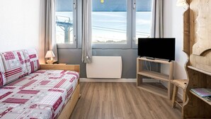 A compact hotel room with a bed, a TV, a wooden shelf, and a window with curtains.