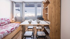 A cozy room with a wooden table, two stools, a sofa with patterned bedding, and a large window with a ski lift view.