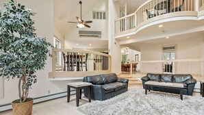 Un vaste espace de vie agrémenté d’un grand arbre en pot, de deux canapés en cuir, d’un tapis moelleux et d’un ventilateur de plafond.