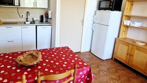 A compact kitchen with white cabinets, a microwave, a small refrigerator, and a wooden cabinet with shelves.