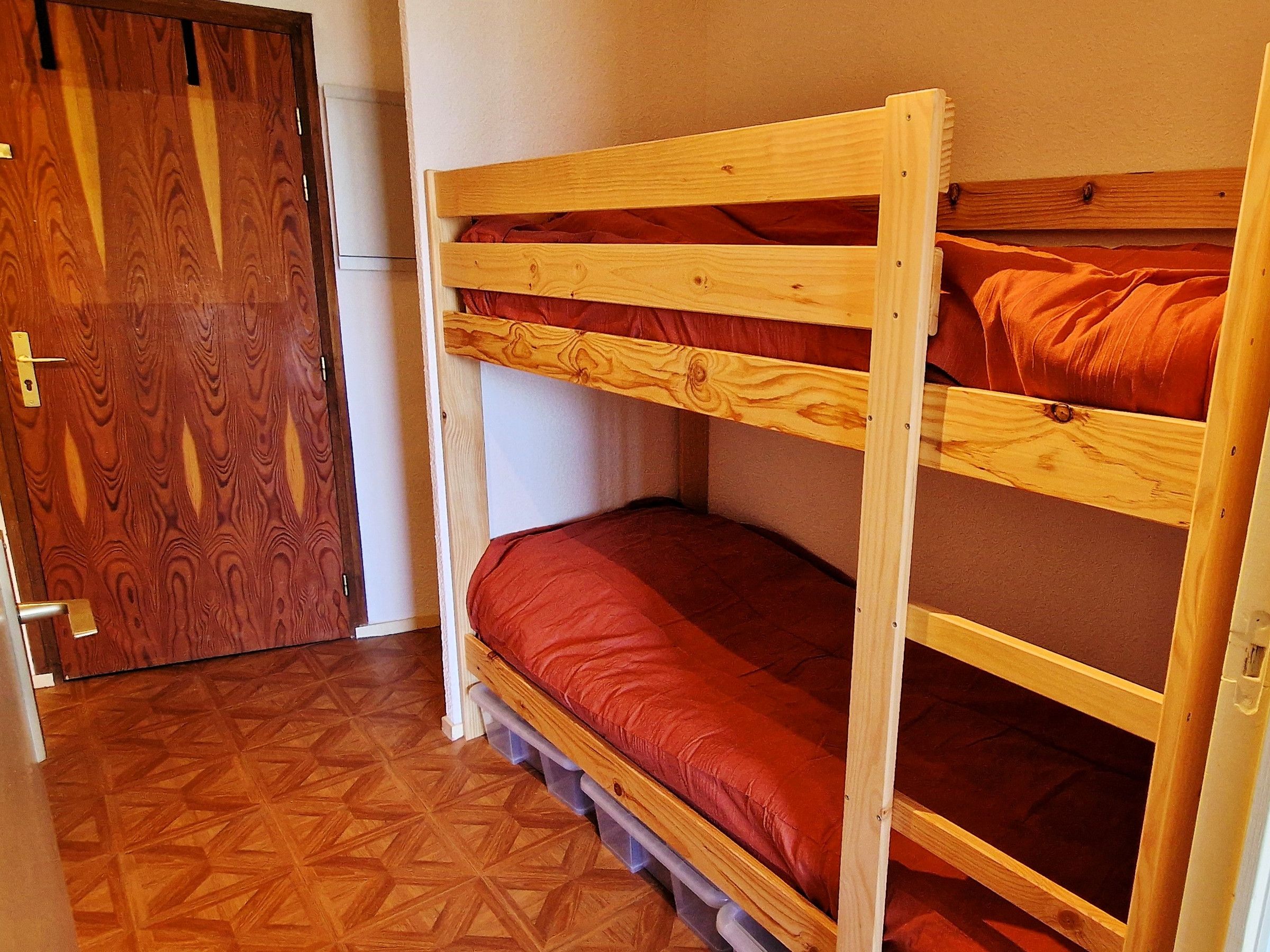 Un lit superposé avec un matelas rouge, dans une pièce dont le sol et la porte sont en bois.