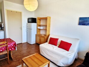 Ein kompakter Wohnraum mit einem weißen Sofa, roten Kissen, einem Holztisch, einem weißen Kühlschrank und einem Holzschrank.