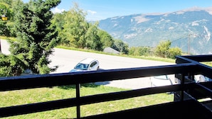 A view from a balcony overlooking a road with a car parked on the side, surrounded by greenery and mountains in the background.
