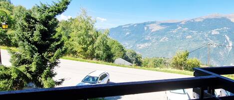 Vista desde un balcón sobre una calle con un coche estacionado al lado, rodeado de vegetación y montañas al fondo.