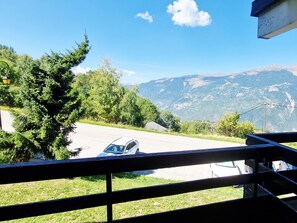 Blick vom Balkon auf eine Straße mit einem parkenden Auto am Rand, umgeben von Grünflächen und Bergen im Hintergrund.