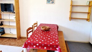 A dining area with a red tablecloth, wooden chairs, a clock, a poster, and a shelf with a bottle.