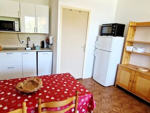 Eine kleine Küche mit weißen Schränken, einer Mikrowelle, einem kleinen Kühlschrank und einem Holzschrank mit Regalen.
