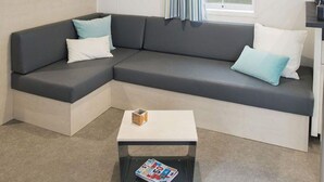 Ein modernes Wohnzimmer mit einem grauen Ecksofa, weißen und blauen Kissen, einem kleinen weißen Couchtisch mit Zeitschriften und einem Fenster mit Vorhängen.