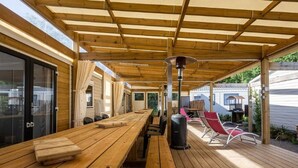 A covered outdoor area with wooden tables and chairs, a grill, and a view of a house in the background. - Mobile home with terrace for 7 people (Bretignolles-sur-Mer)