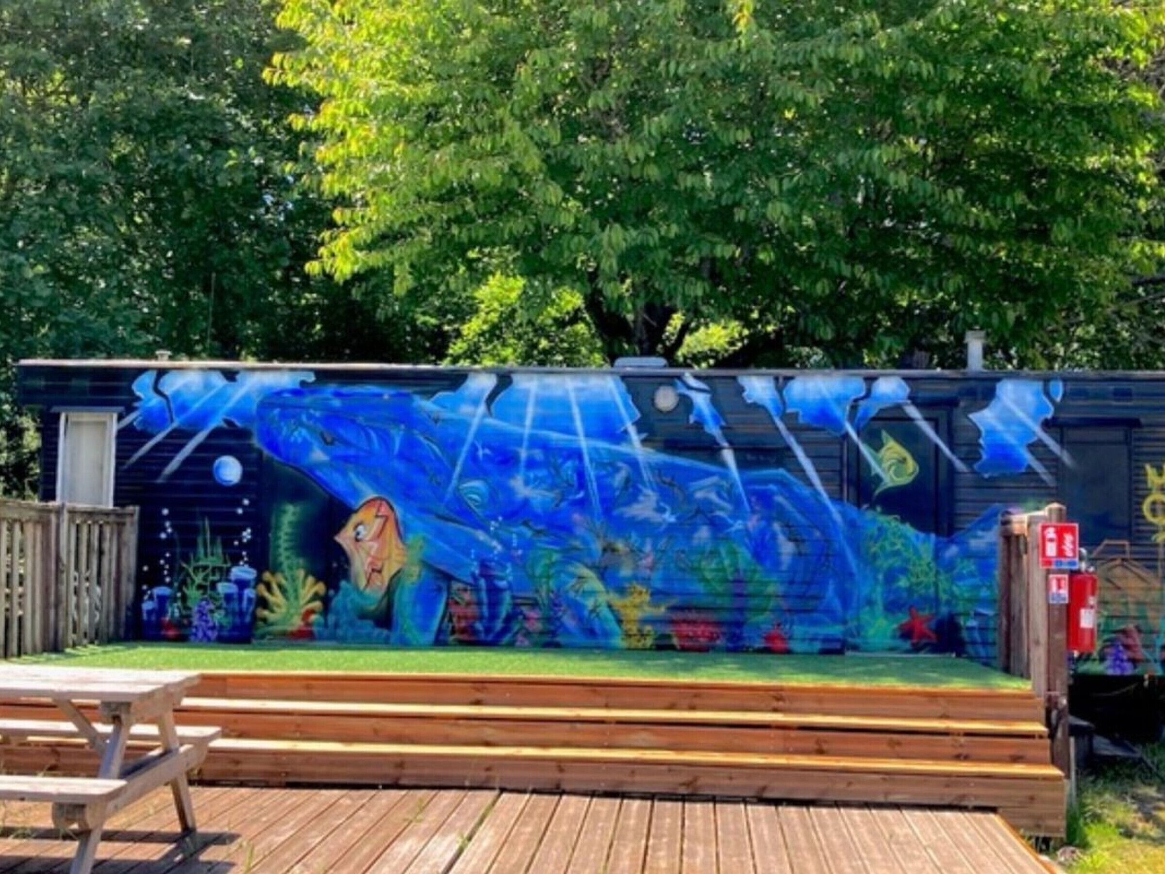 A wooden deck with a mural of a blue ocean scene, a picnic table, and a small building with a door.
