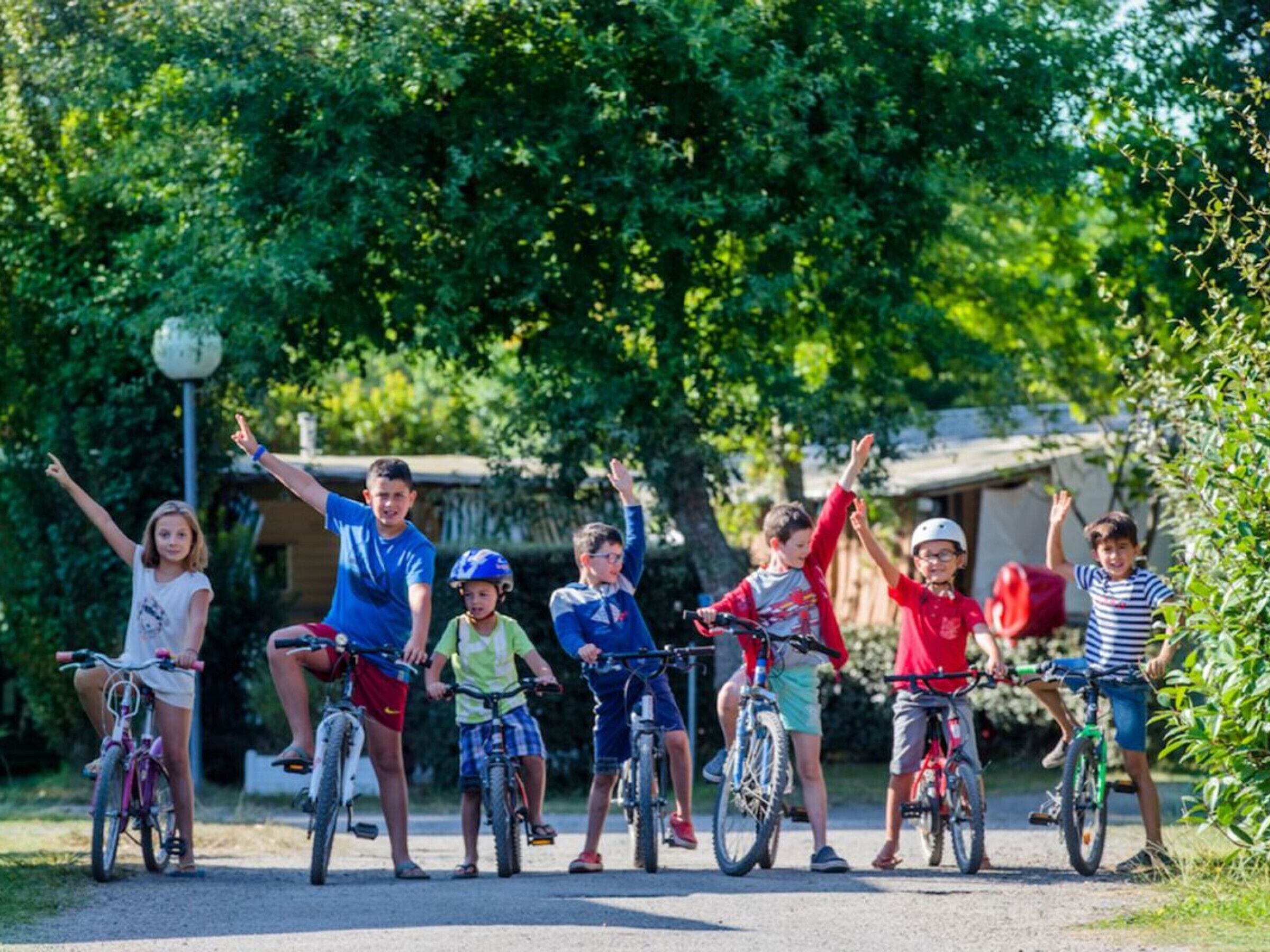 Um grupo de crianças em bicicletas num espaço ao ar livre, com vegetação e um edifício ao fundo.