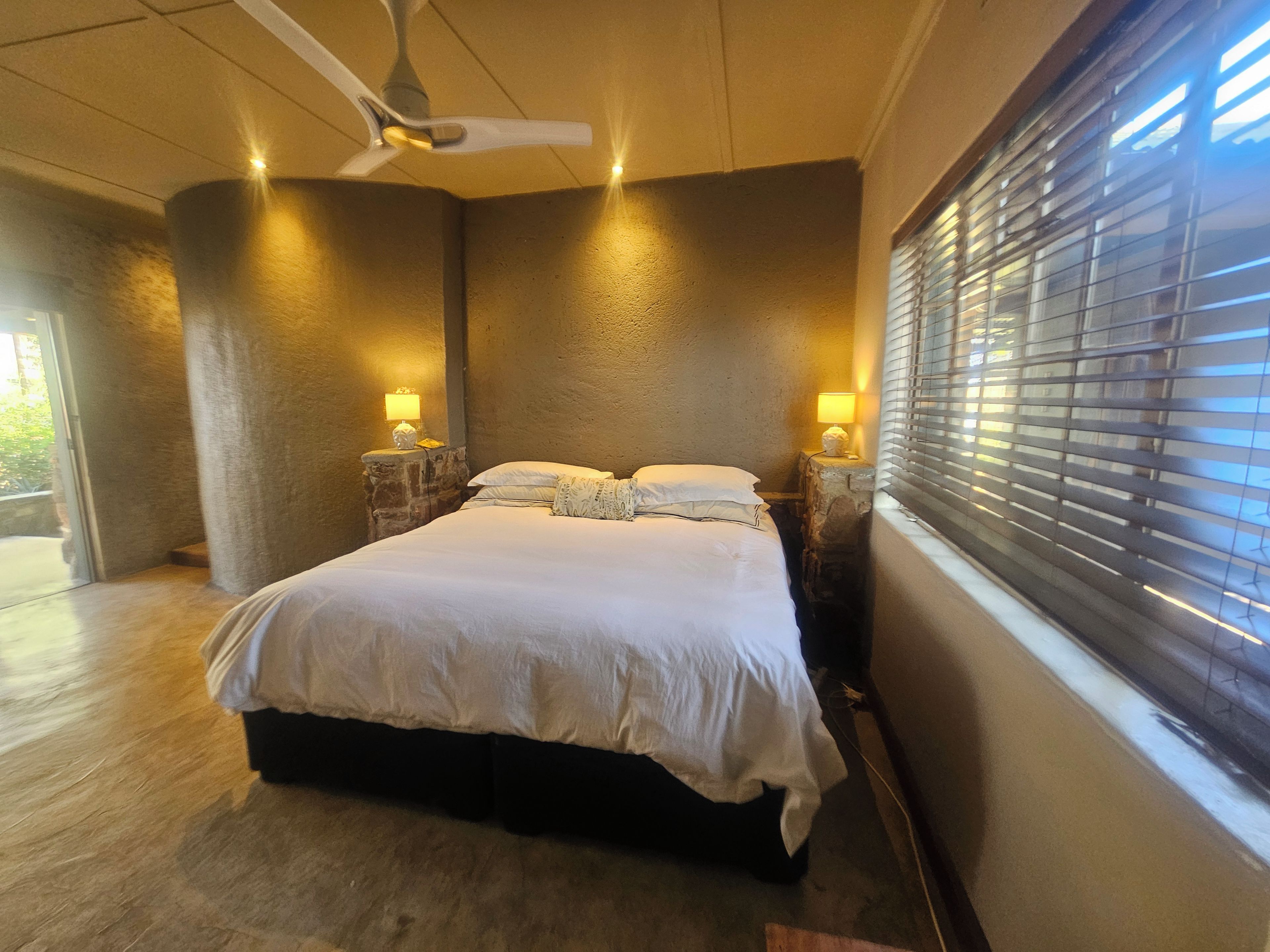 Une chambre à coucher avec un lit, deux lampes de chevet, un ventilateur de plafond et une fenêtre avec des stores.