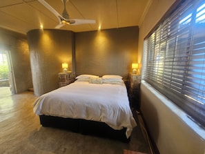 Quarto com cama, duas luminárias de cabeceira, ventilador de teto e janela com persianas.