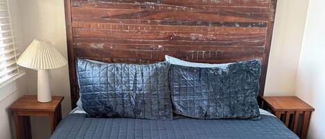 Una cama con cabecera de madera, dos almohadas y una colcha azul con textura.