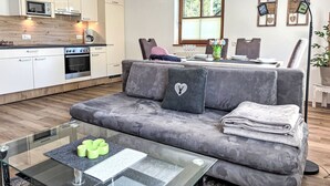 Eine moderne Küche mit weißen Schränken, einer Mikrowelle und einem Herd. Es gibt ein graues Sofa mit einem herzförmigen Kissen und einen Glastisch mit einem grünen Kerzenhalter.