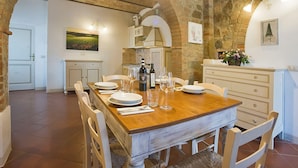 Un coin repas avec une table en bois dressée pour quatre personnes, des chaises blanches, un mur en briques, un luminaire suspendu et une cuisine en arrière-plan.