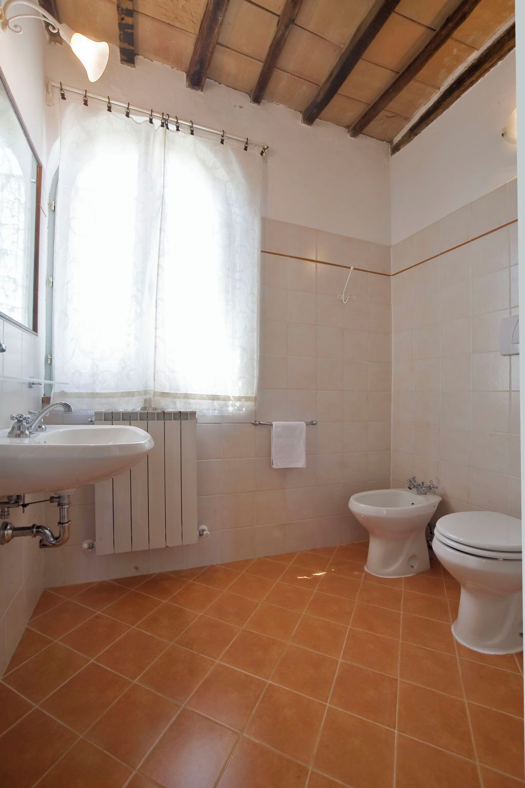 Une salle de bain avec un lavabo, deux toilettes, une fenêtre avec un rideau et un sol carrelé.