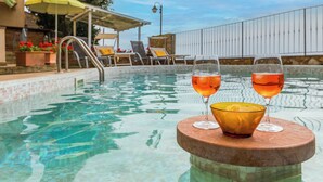 A poolside setting with two glasses of a beverage on a round table, a pool, and lounge chairs in the background.