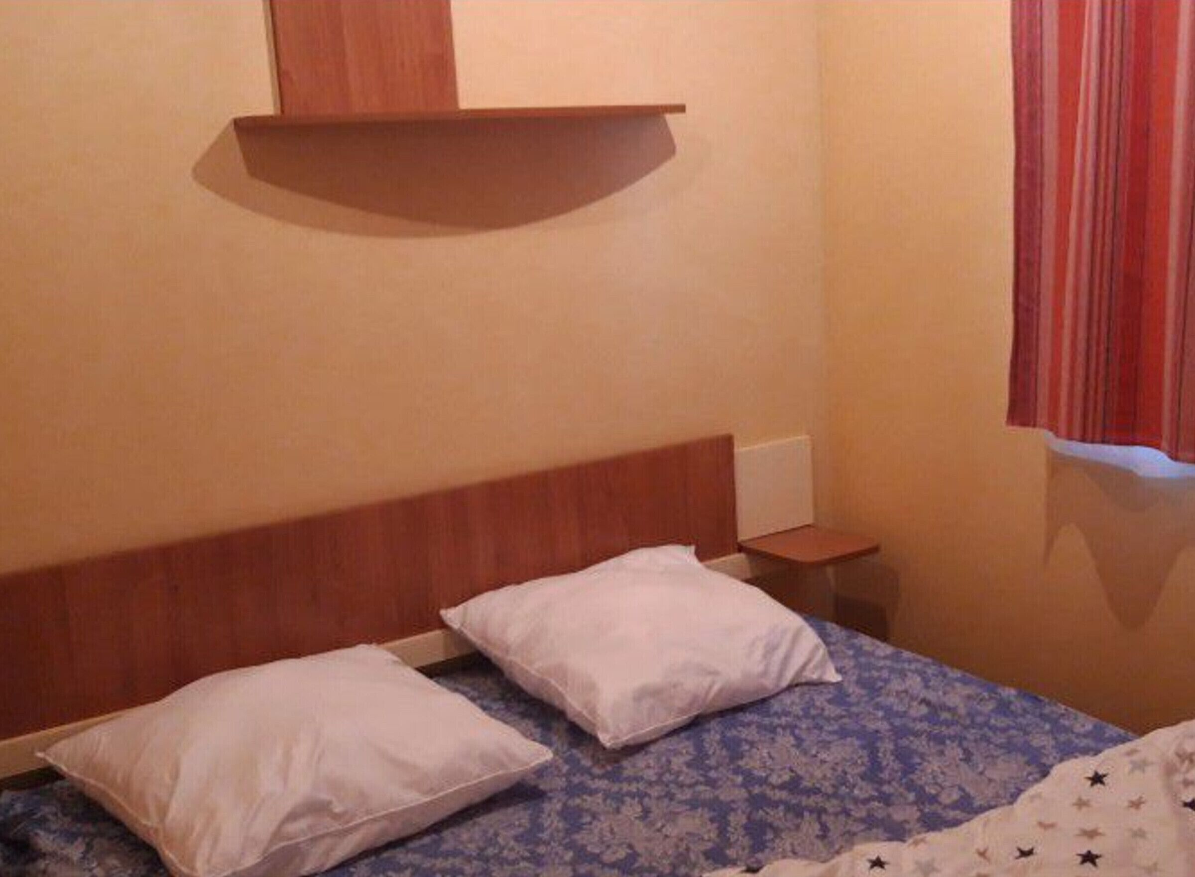 Una habitación pequeña y sencilla con una cama, dos almohadas, una mesita de noche y una cortina a rayas rojas.