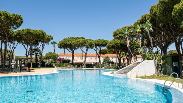A large swimming pool with a curved edge, surrounded by trees and a building with a red-tiled roof.