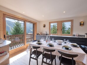 Un comedor con mesa preparada para cuatro personas, ventanas amplias con vistas a la montaña y una terraza de madera.