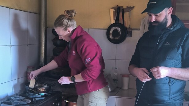 Une cuisine avec un cuisinière à gaz, une femme qui cuisine et un homme qui se tient à proximité.